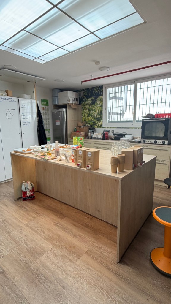 A modern kitchen with a wooden island, featuring various food items and drink containers on top. A fridge and appliances are visible in the background, along with a large window letting in natural light.